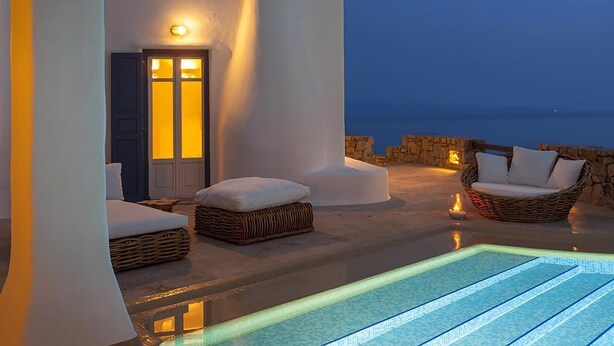 A modern outdoor pool area with white walls and wicker furniture, overlooking a stone wall and the sea.