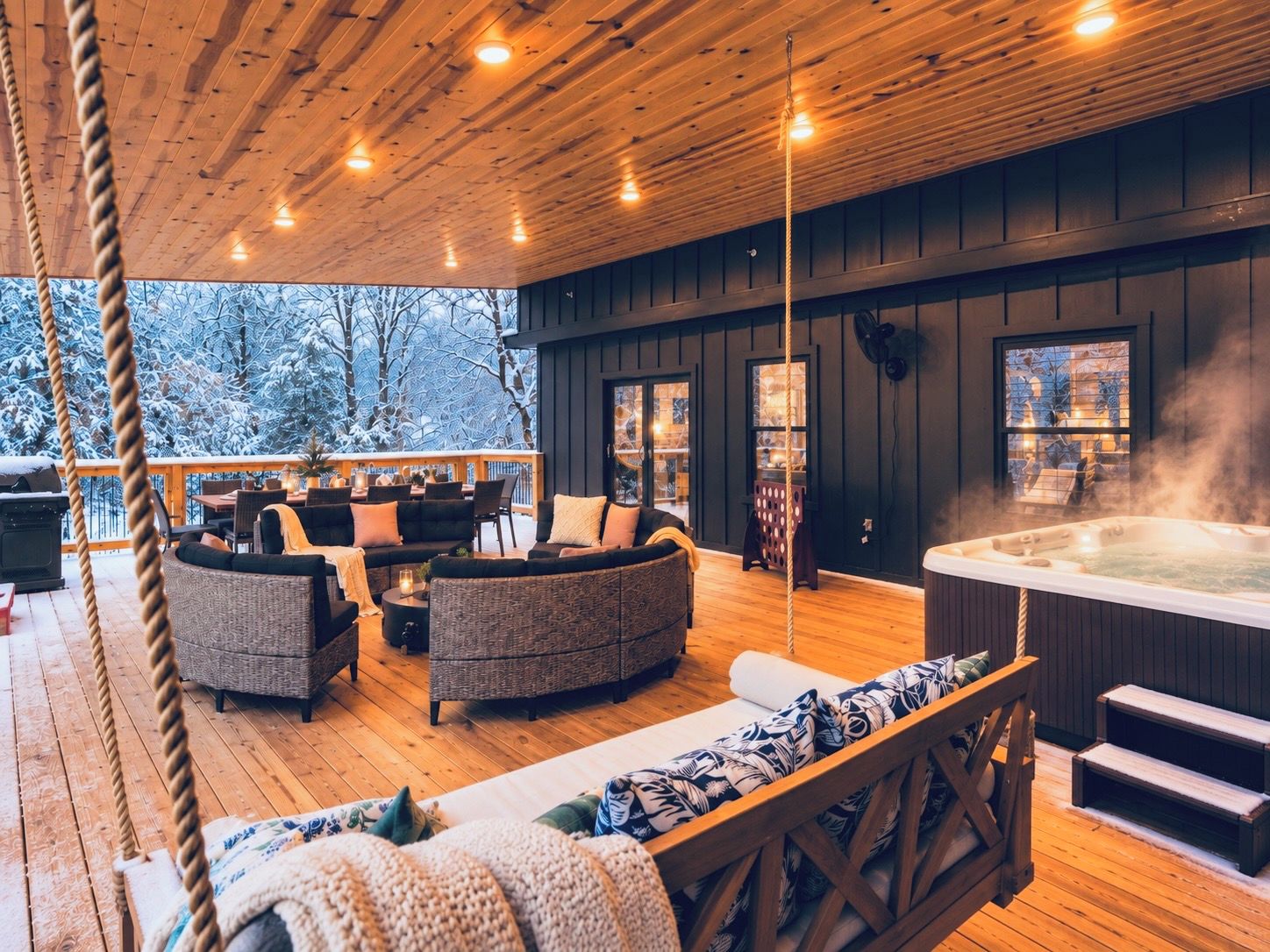 A cozy outdoor seating area with a hot tub, wooden deck, and a view of snow-covered trees.