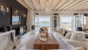 A modern living room with a wooden ceiling, a large window offering a sea view, a white sofa, a wooden coffee table, and a chandelier.