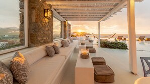 A sunlit outdoor seating area with stone walls, a long sofa, and a view of the sunset over the sea.