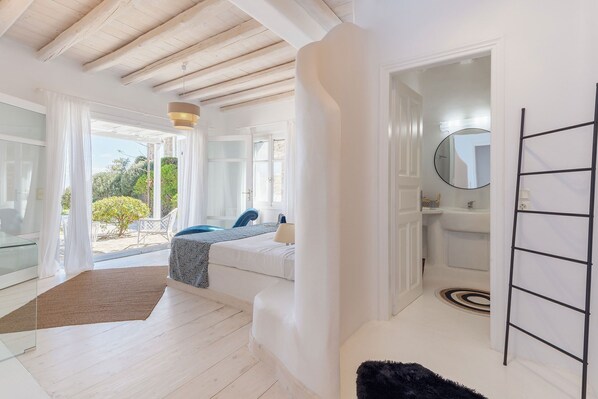 A bright room with a bed, a small table, and a chair. There is a window with curtains, a door leading to a bathroom, and a view of greenery outside.
