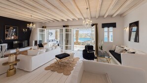 A spacious living room with a large white sofa, a black armchair, and a wooden coffee table. There is a TV on the left and a view of the sea through the open door.