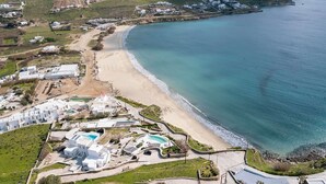 An aerial view of a coastal resort with a sandy beach, clear blue waters, and white buildings with a pool area.