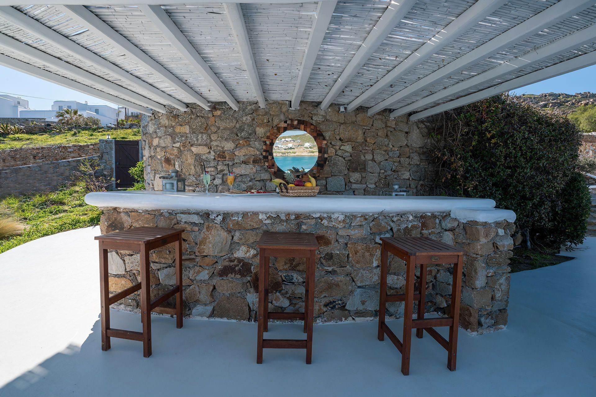 Zona de bar al aire libre con pared de piedra, mostrador de madera y tres taburetes de madera.