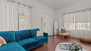 A modern living room with a blue sofa, a coffee table with magazines, a floor lamp, a mirror, and a view of the outdoors through large windows.