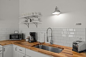 Une cuisine moderne avec un plan de travail en bois, un évier en acier inoxydable, une machine à café, un grille-pain et une étagère murale où sont posées des tasses et une horloge.