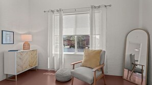 A room with a window, a chair, a side table, a lamp, a mirror, and a framed certificate on the wall.
