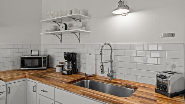A modern kitchen with a wooden countertop, stainless steel sink, a coffee maker, a toaster, and a wall-mounted shelf with cups and a clock.