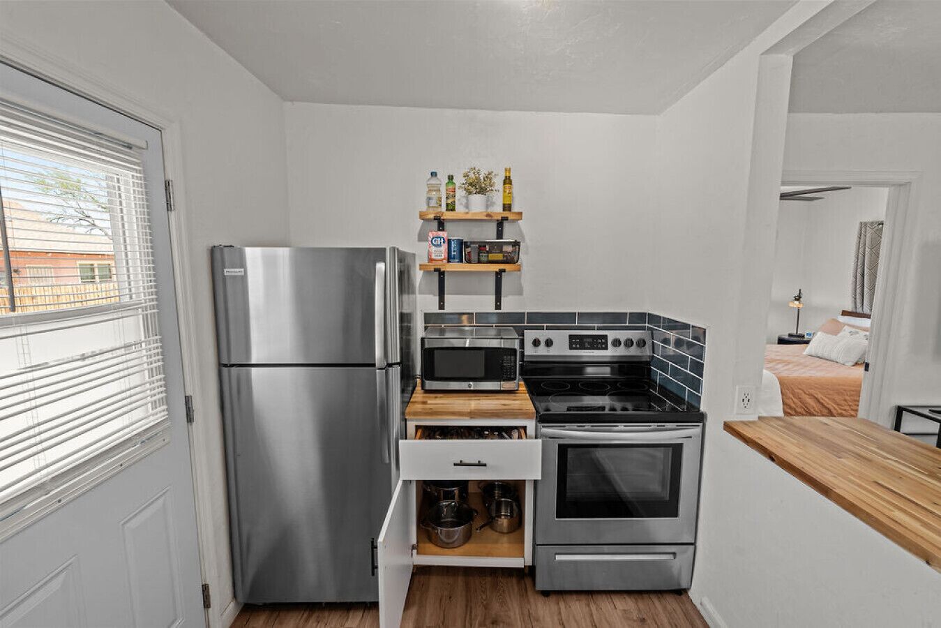 A compact kitchen with a refrigerator, microwave, stove, and oven. There is a wooden countertop and a shelf with bottles and containers.