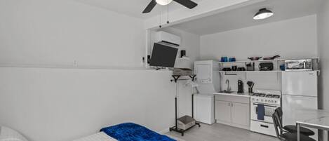 A compact room with a bed, a desk, a kitchenette, and a ceiling fan.