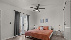 Un dormitorio con una cama, un ventilador de techo, una mesita de noche, una lámpara y una puerta que da a un balcón.