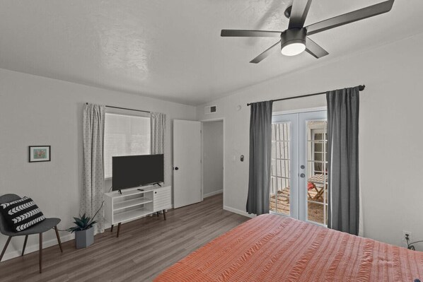 Un dormitorio moderno con una televisión de pantalla plana sobre un mueble blanco, un ventilador de techo, una cama con colcha naranja, una silla con cojín blanco y negro, y una planta en maceta.