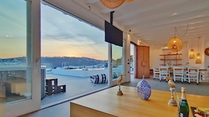 A modern interior with large windows, a dining area with white chairs, and decorative items on the table. - My Rental Homes - Villa Waves with pool and Jacuzzi. Close to Platis Gialos beach (Mykonos)