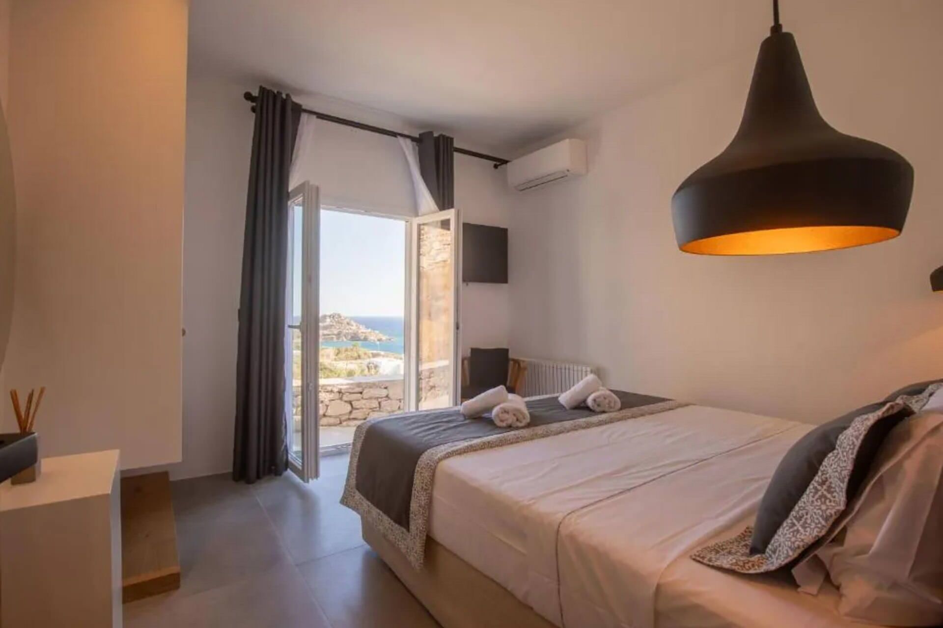 A bedroom with a bed, a hanging lamp, a view of the sea, and an air conditioning unit.