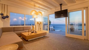 A modern living room with a large window overlooking the sea, a sofa, a coffee table, and a flat-screen TV mounted on the wall. - My Rental Homes - Villa Waves with pool and Jacuzzi. Close to Platis Gialos beach (Mykonos)