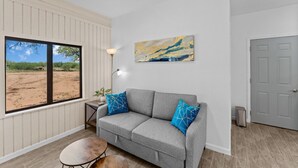 Una sala moderna con un sofá gris, dos cojines azules estampados, una pequeña mesa redonda, una lámpara de pie y un amplio ventanal con vistas a un paisaje desértico.