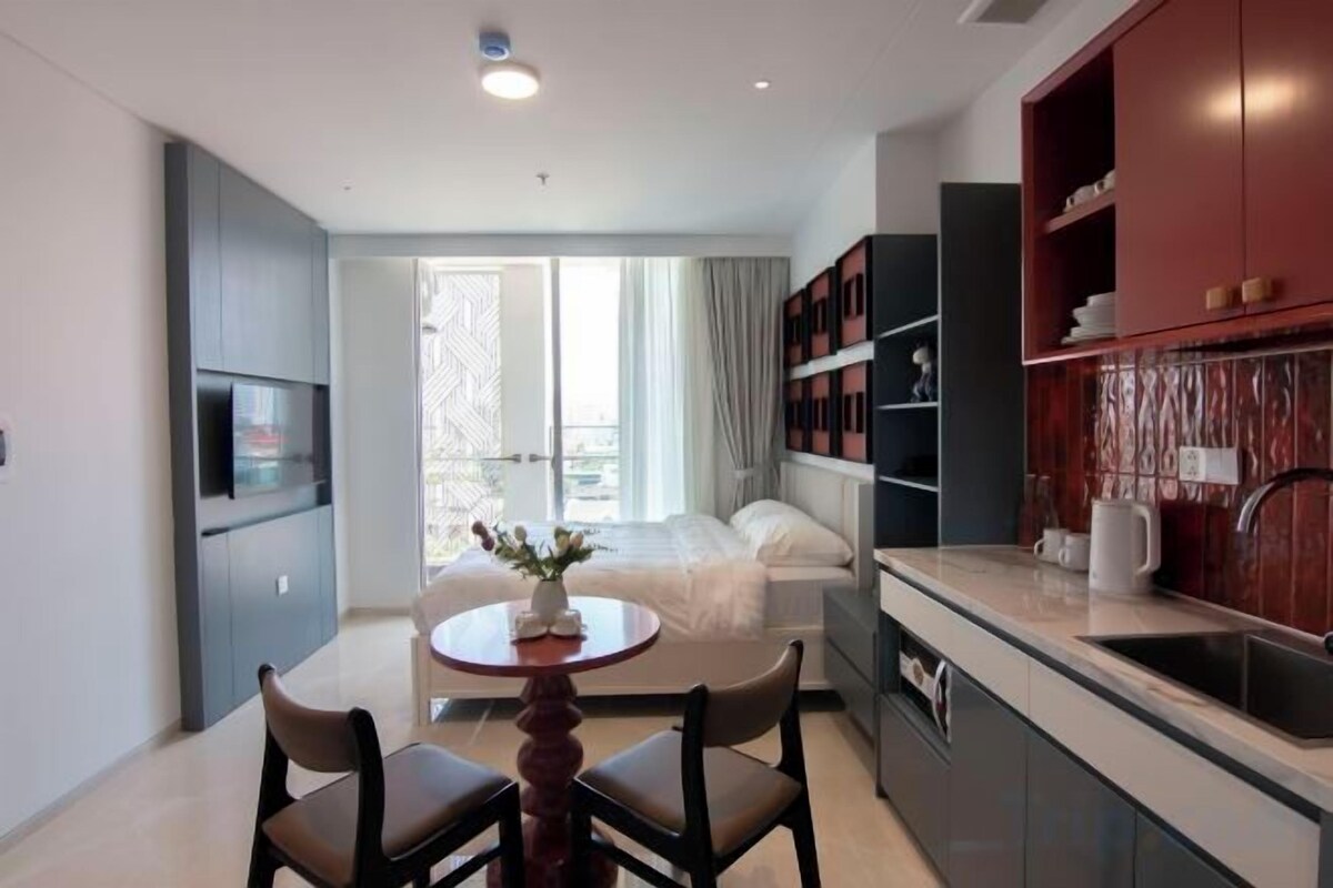 A compact hotel room with a bed, a small dining table with chairs, a kitchenette, and a TV.