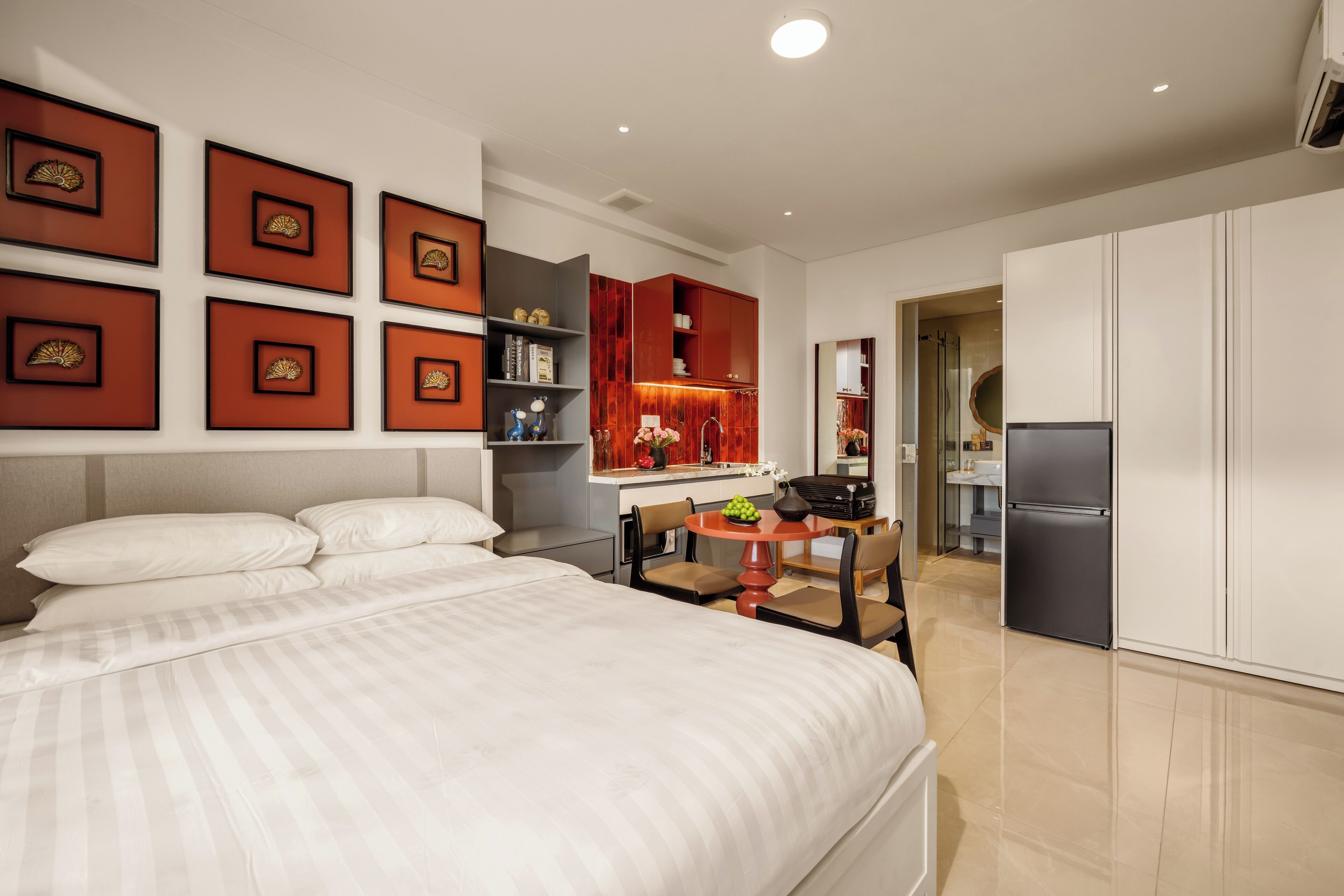 A modern hotel room with a large bed, a dining area with a table and chairs, a kitchen with a refrigerator, and a bathroom visible through an open door.