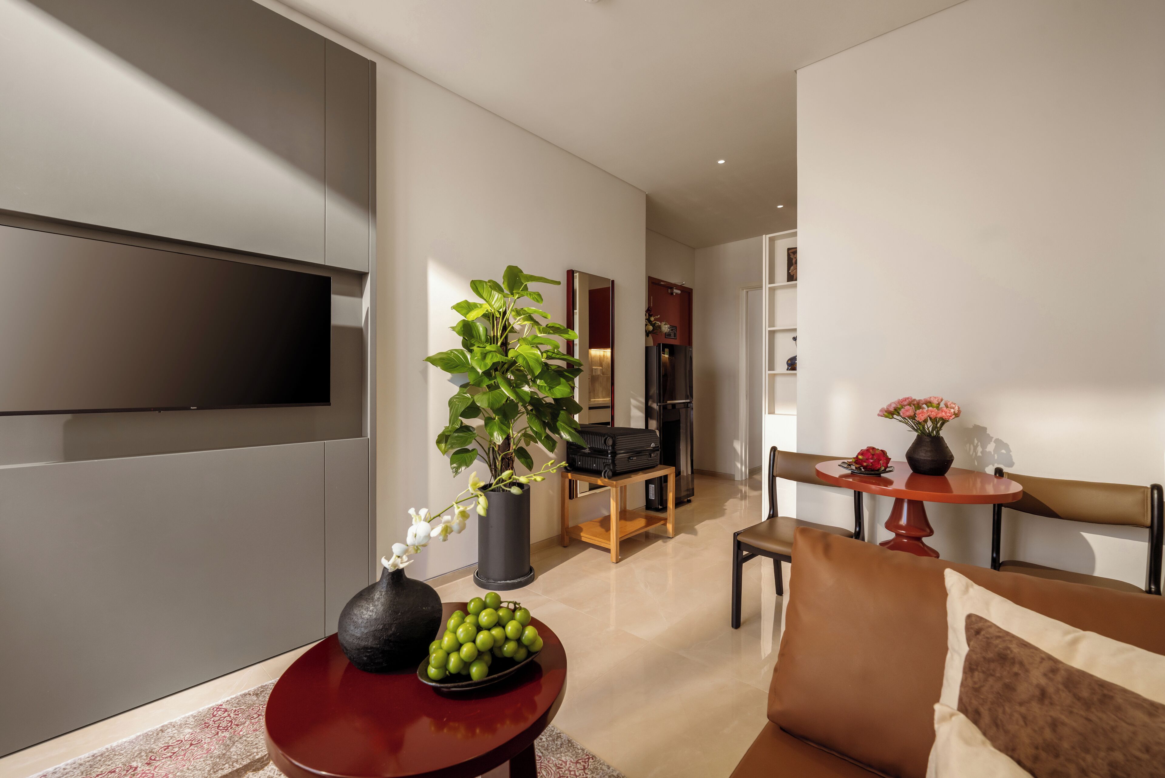 A modern living room with a flat-screen TV, a potted plant, a bowl of fruit, and a dining area with a table and chairs.