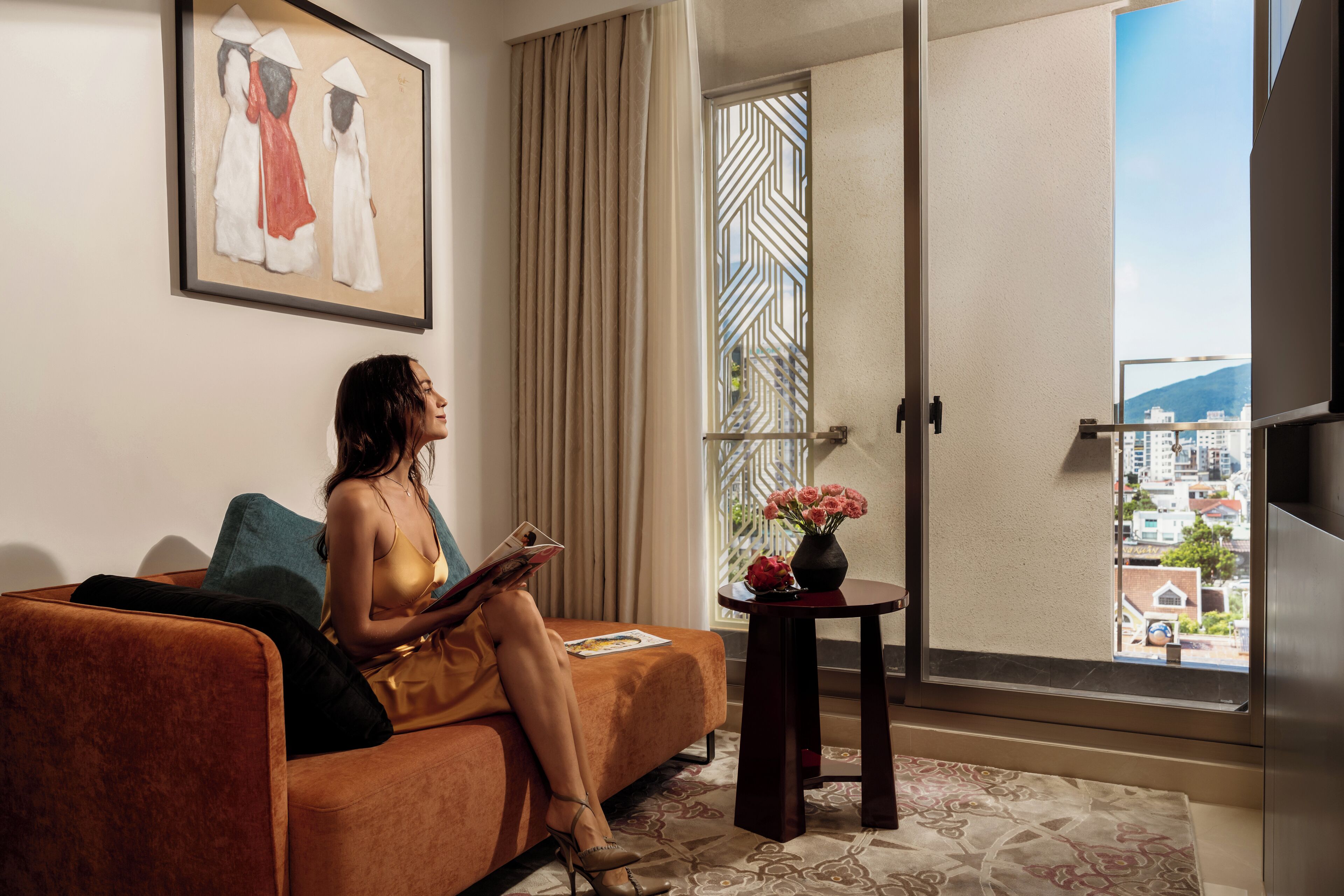A woman sitting on a sofa reading a magazine in a room with a large window, a painting on the wall, and a small table with flowers.