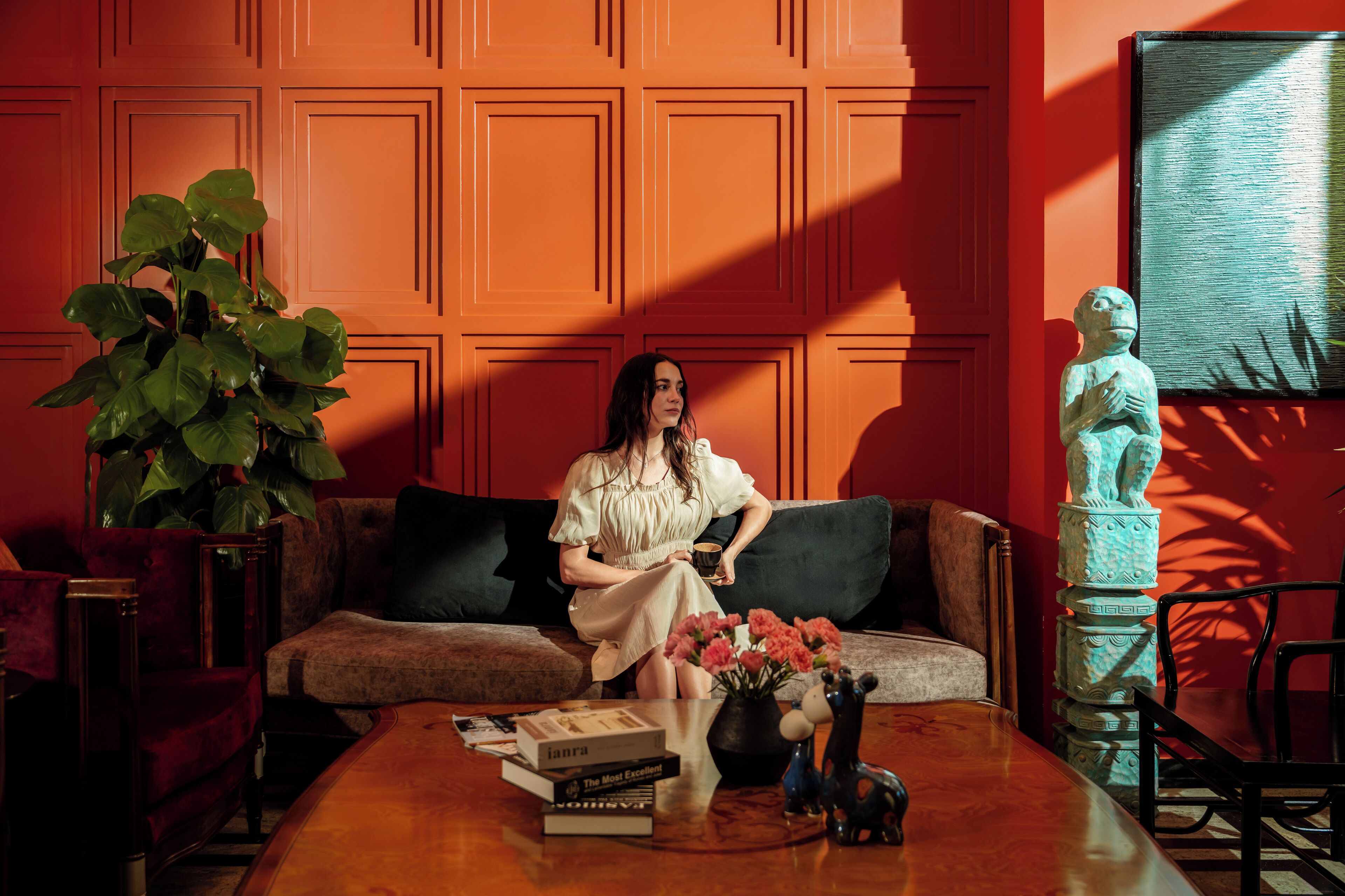 Una habitación con un muro empapelado de color rojo, un sofá, una mesa de centro con libros y un jarrón, y una estatua.