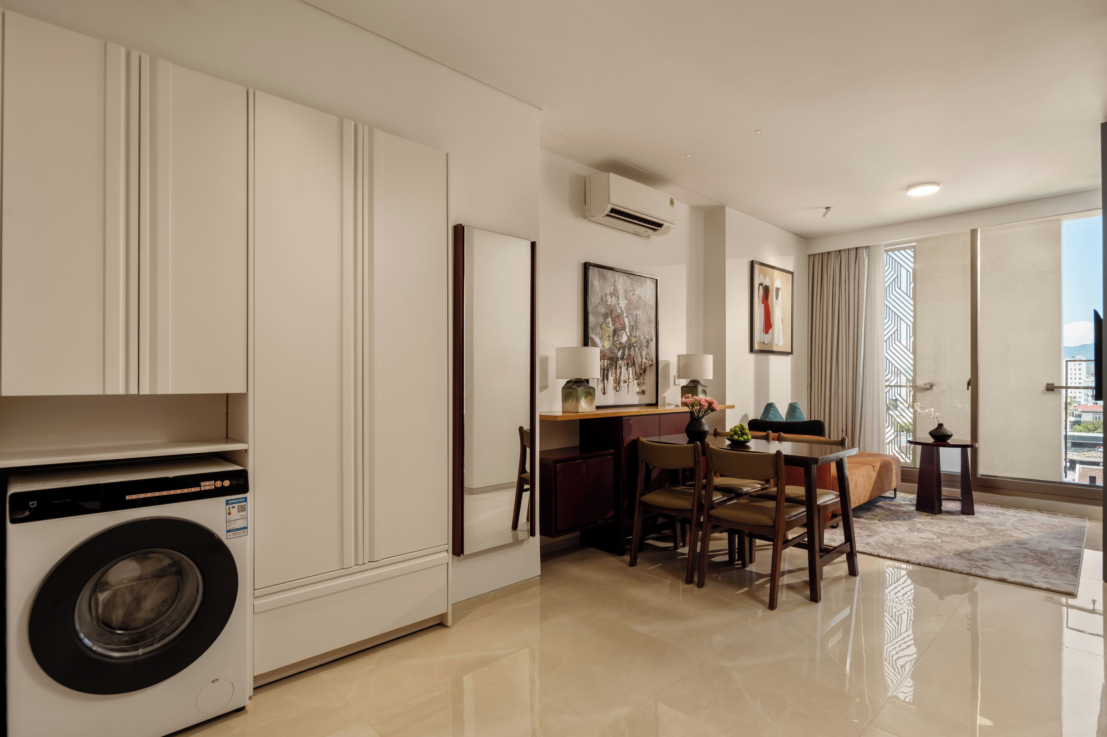 A modern hotel room with a washing machine, a dining area with a table and chairs, and large windows with curtains.