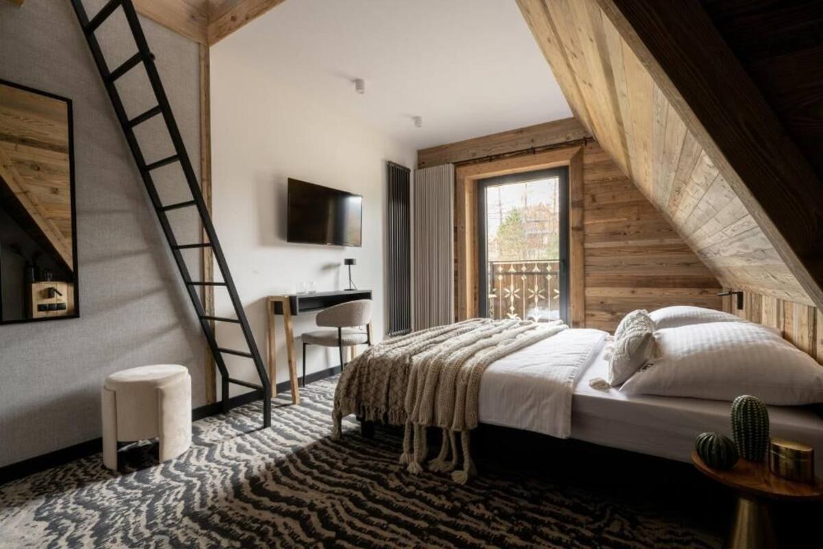 Ein Schlafzimmer mit schrägen Wänden, einem Bett mit Kissen, einem an der Wand befestigten Fernseher, einem Schreibtisch mit Stuhl und einer Leitern.
