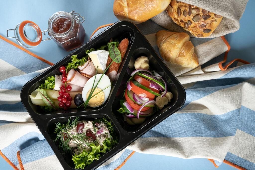 A meal with various food items in a black container, a jar of sauce, and a loaf of bread on a striped cloth.