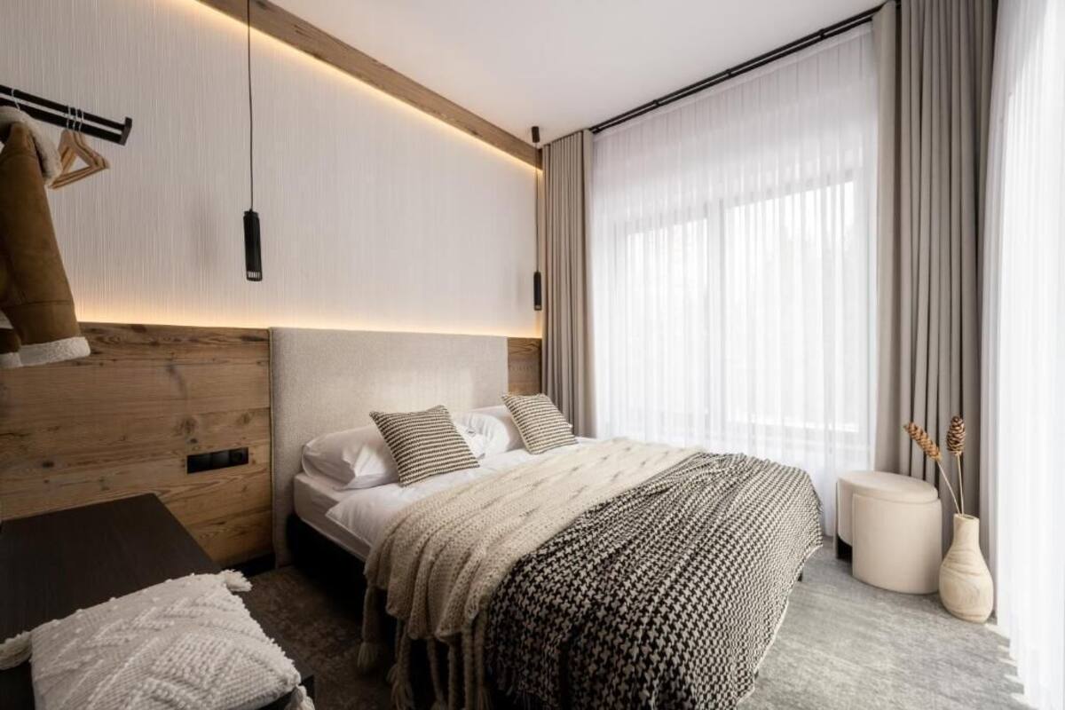 Ein modernes Hotelzimmer mit einem hölzernen Kopfteil, einem großen Bett mit vielen Kissen, einem Nachttisch, einem kleinen Hocker und einer Vase mit Trockenpflanzen.