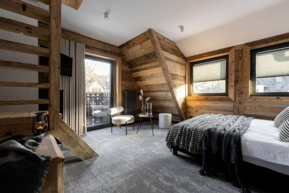 Ein Schlafzimmer mit schräg versiegelter Decke, Holzbalken, einem Bett mit gemustertem Überwurf, einem Sessel, einem kleinen Tisch mit einer Vase und einem Fenster mit Jalousien.