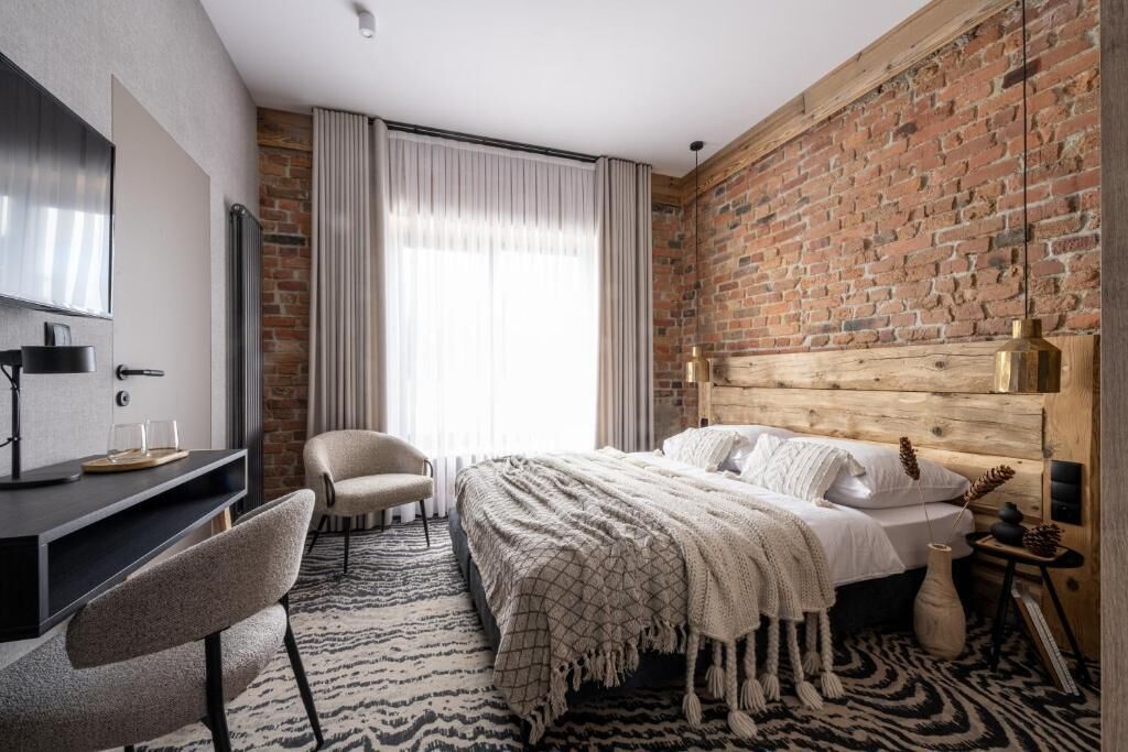 A modern bedroom with a brick wall, a wooden headboard, a bed with white linens, a desk with a chair, and a large window with curtains.