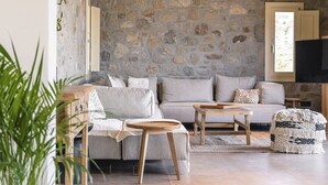 A cozy living room with a stone wall, a grey sofa, a wooden coffee table, and a patterned ottoman. - My Rental Homes - Villa Wolf with private pool and sea view (Mykonos)