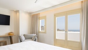 A hotel room with a large bed, a chair, a small table, and a TV mounted on the wall. There are two windows with curtains, and a view of the sea is visible through the windows. - My Rental Homes - Villa Wolf with private pool and sea view (Mykonos)