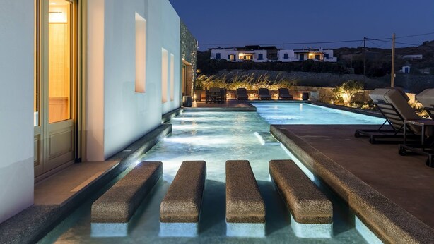 A pool area with a water feature, lounge chairs, and a building with a lighted entrance. - My Rental Homes - Villa Wolf with private pool and sea view (Mykonos)