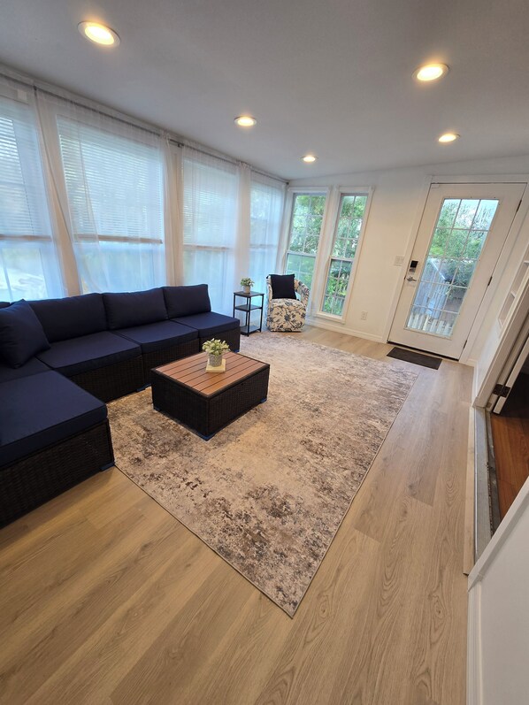 A living room with a sectional sofa, a coffee table, and a chair. There are large windows and a door leading outside.