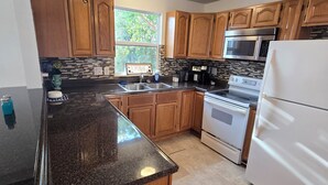 A kitchen with wooden cabinets, a stainless steel microwave, a white refrigerator, a granite countertop, and a window with a view of greenery. - Spacious 5-bedroom beautiful water view home with private boat dock! (Homosassa)