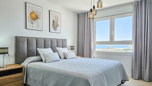 A modern bedroom with a large bed, grey headboard, and a view of the city through the window. - 4 bedroom townhouse in La Cala (Mijas Costa)