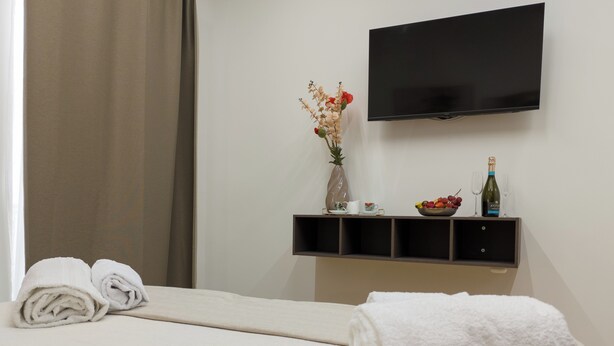 Hotellihuone, jossa on sänky, seinälle asennettu televisio, maljakko kukkineen hyllyllä, pullo viiniä ja kulho täynnä hedelmiä.