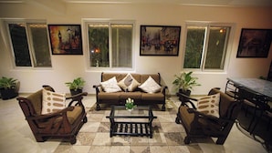 A living room with a sofa set, coffee table, and two armchairs. There are potted plants, a dining table with chairs, and artwork on the walls.