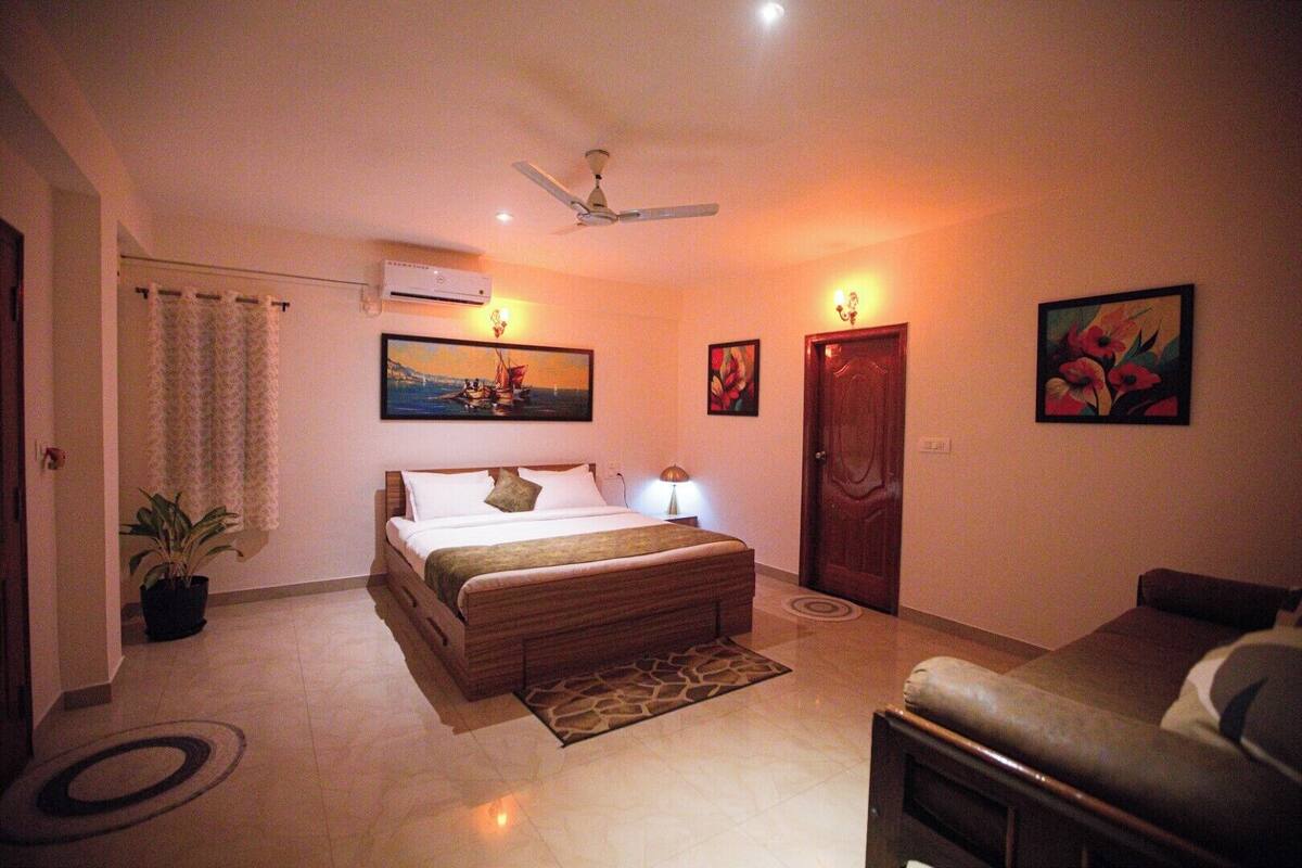 A bedroom with a bed, a ceiling fan, a potted plant, a nightstand, a lamp, a door, and a painting on the wall.