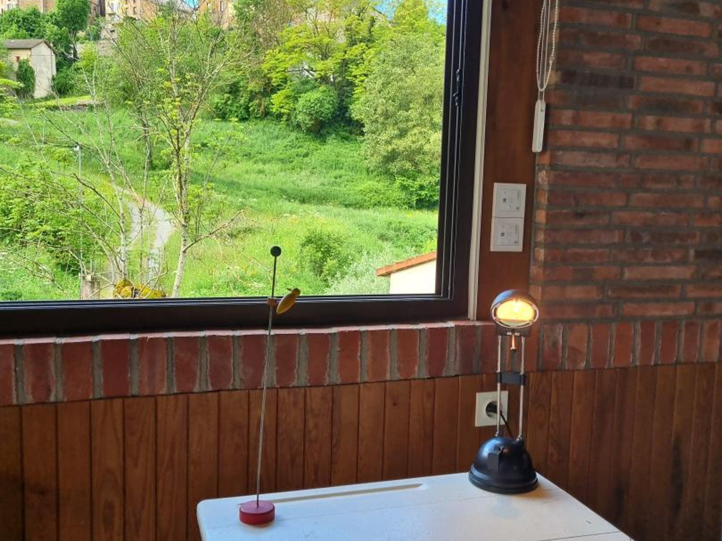 Una stanza con una finestra che si affaccia su un paesaggio verde, un tavolino e un punto luce a parete.