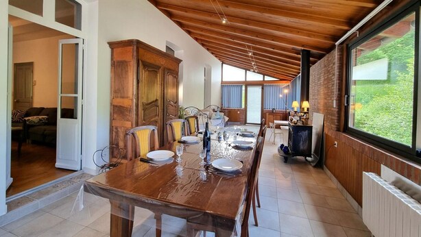 A dining area with a wooden table set for six, flanked by chairs, a cabinet, and a view of the outdoors through large windows.