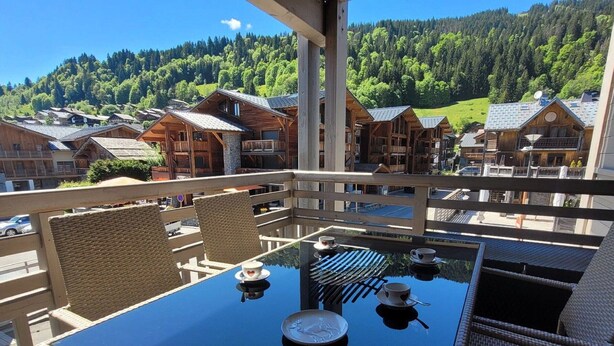 A balcony with wicker chairs and a glass table, overlooking a village with wooden houses and a forested hill in the background. - Chic apartment in Les Gets center, sleeps 6, near ski and shops (Les Gets)