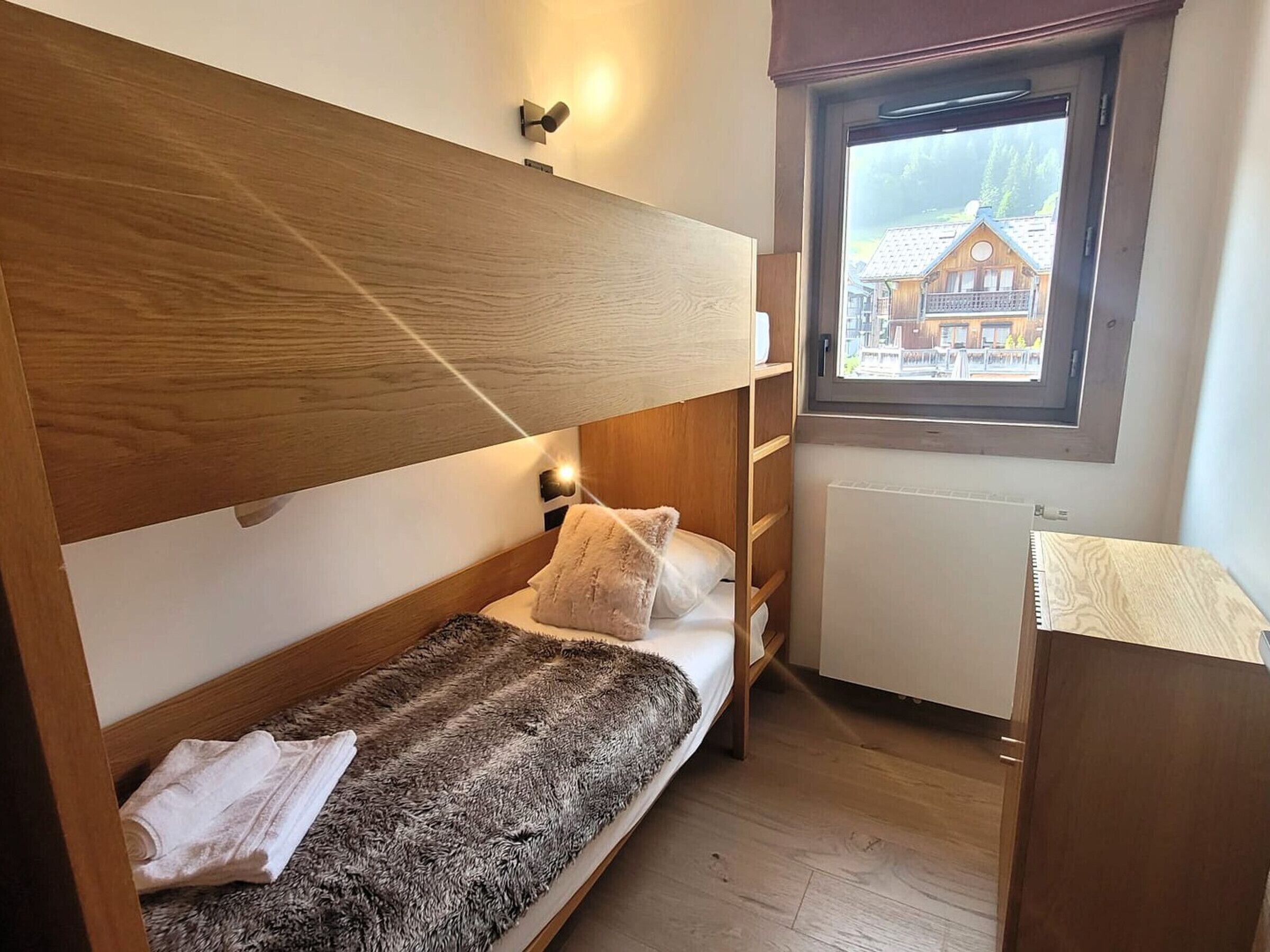 A compact bedroom with a bunk bed, a small desk, and a window with a view of a house.