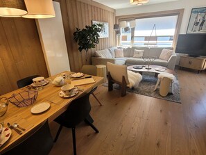 Un salon chaleureux avec un ensemble de canapés, un coin repas avec une table dressée pour deux personnes, et une grande fenêtre offrant une vue agréable.