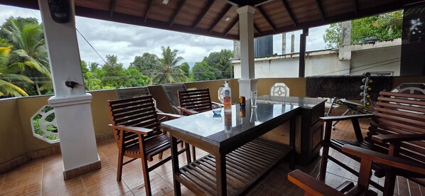 A covered outdoor seating area with a table and chairs, a decorative pillar, and a view of greenery and buildings in the background.