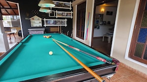 A pool table with balls and cues, a white chair, and a shelf with decorative items. - Kandy Comforts (Katugastota)