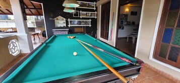 Una mesa de billar con bolas y tacos, una silla blanca y una estantería con adornos.