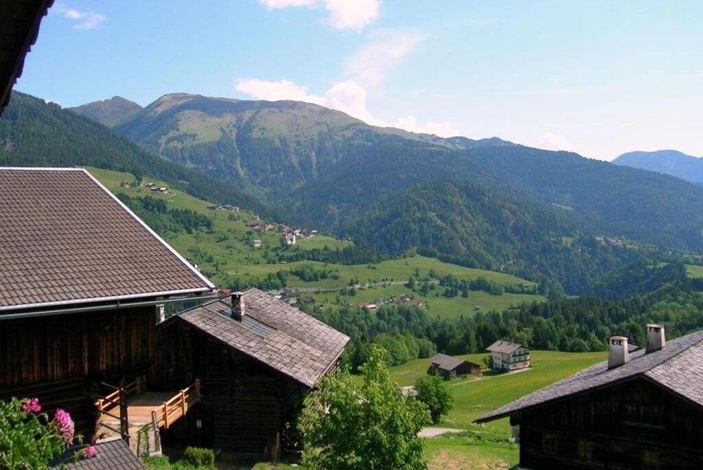 Une cabane en bois, dotée d’un toit en pente, entourée de collines verdoyantes et avec des montagnes en arrière-plan.