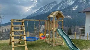 Un parque infantil de madera con tobogán y columpios, con montañas al fondo.